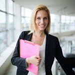 Successful smiling business woman at work holding documents, standing near window in office
