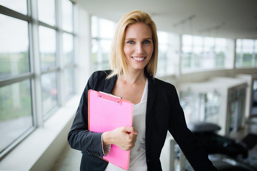 Successful smiling business woman at work holding documents, standing near window in office