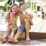 Portrait of little girl and boy with popsicles wearing mirrored sunglasses in front of swimming pool